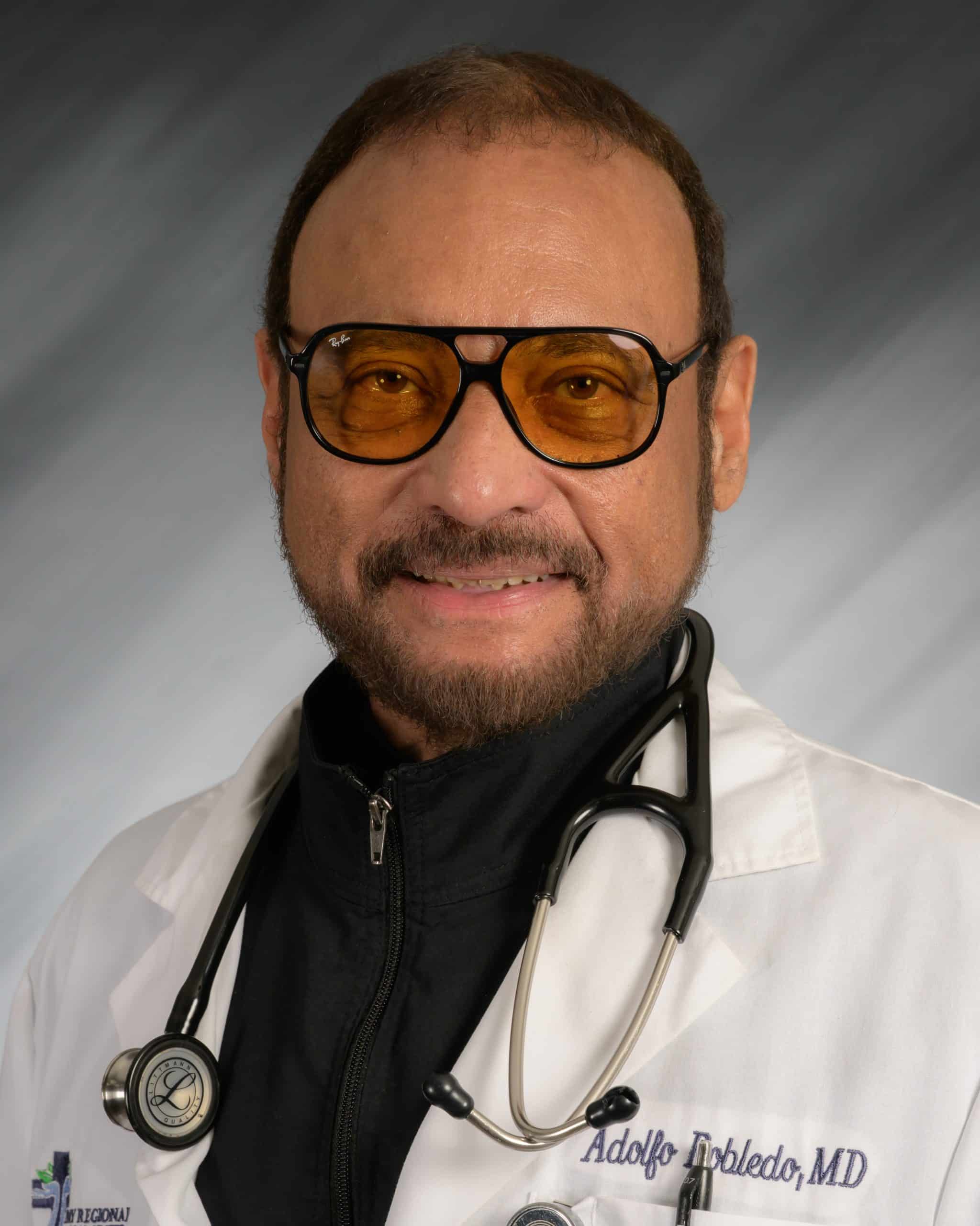Adolfo Toledo, MD, in a white coat with stethoscope, wearing glasses, smiling against a gray background.