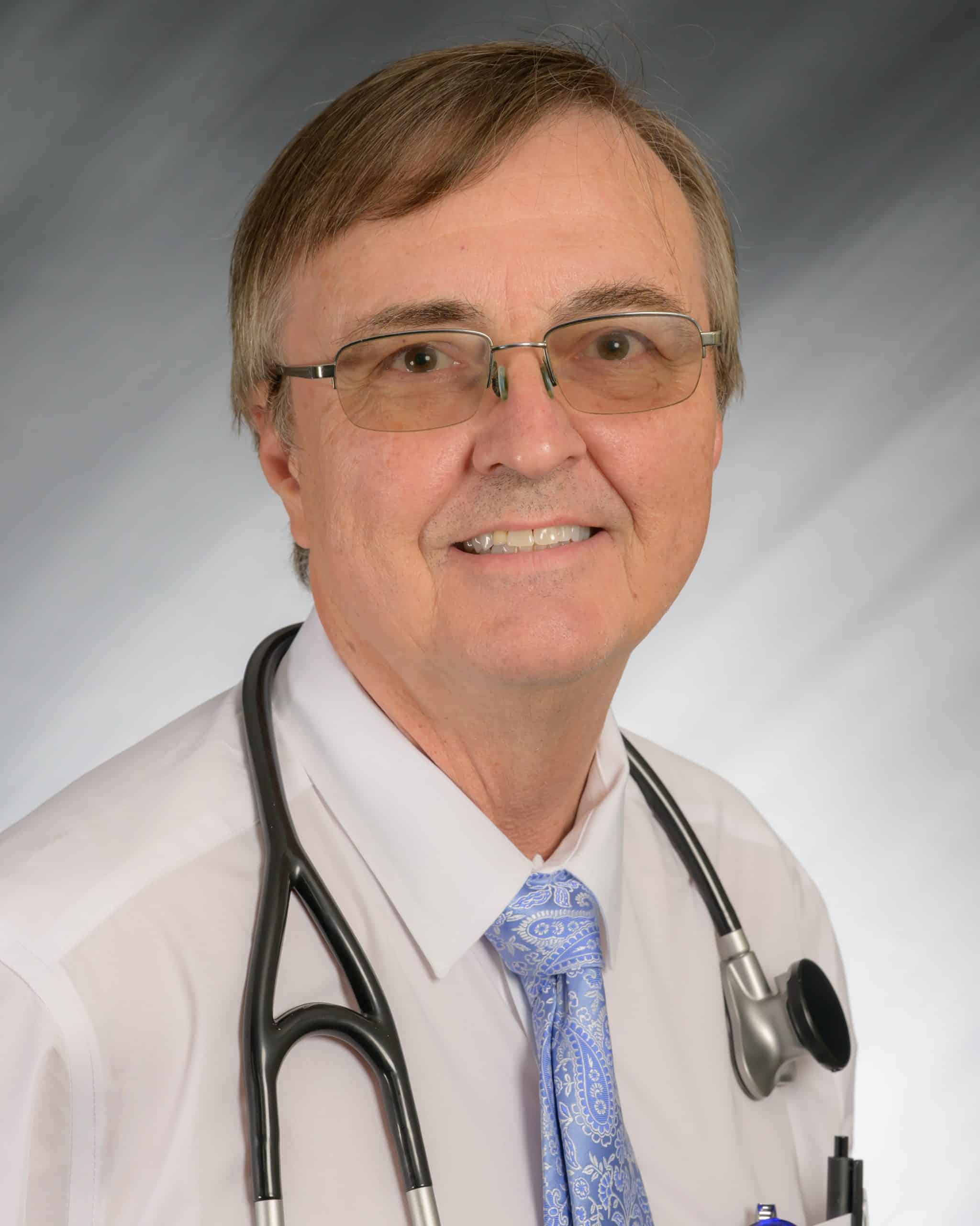 A man with glasses and stethoscope, wearing a white shirt and blue tie.
