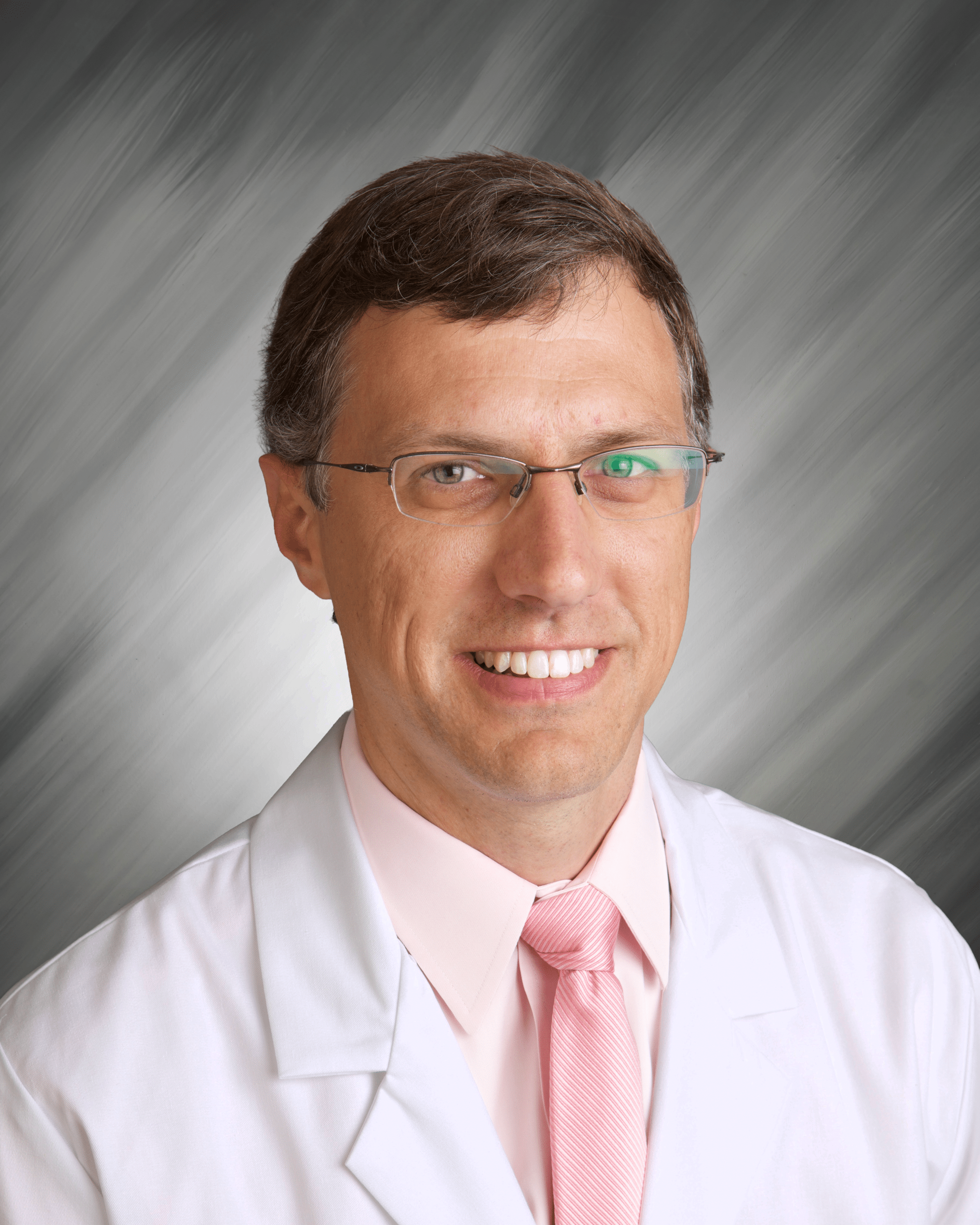 A person in a white coat with glasses and a pink tie smiles at the camera.