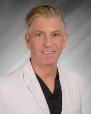 A person with short hair in a white blazer smiling against a gray background.