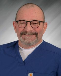 A man with glasses and a beard wearing blue scrubs, smiling against a gray background.