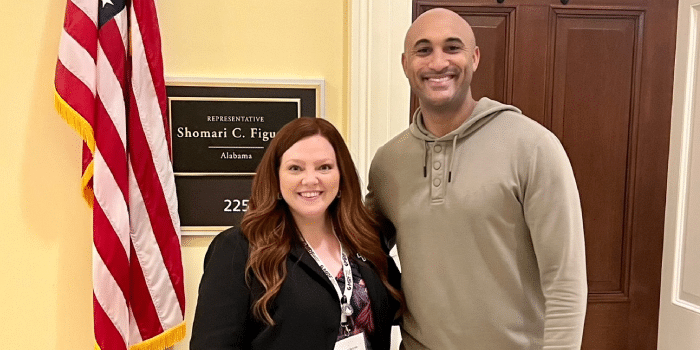 A smiling man and woman standing by a sign for Representative Shomari C. Figures in an office hallway.
