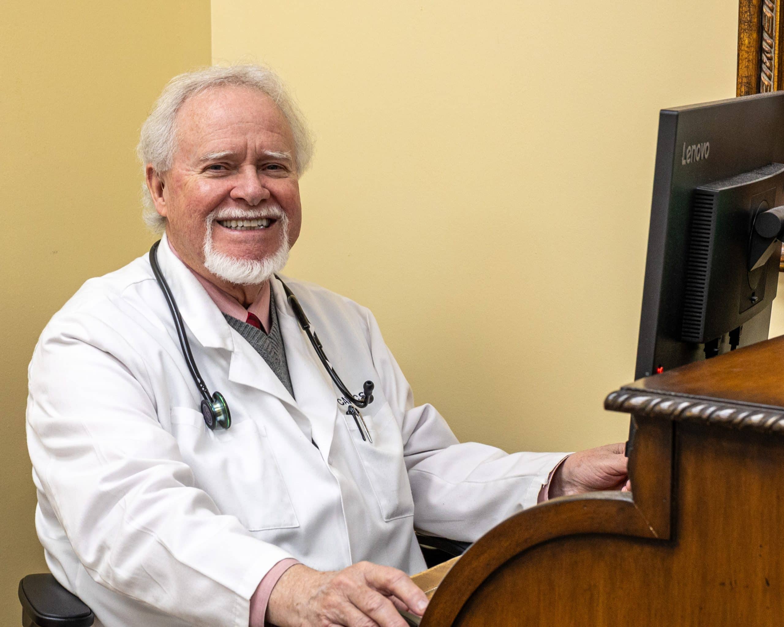 A man in a lab coat with a stethoscope sitting at a desk, smiling.