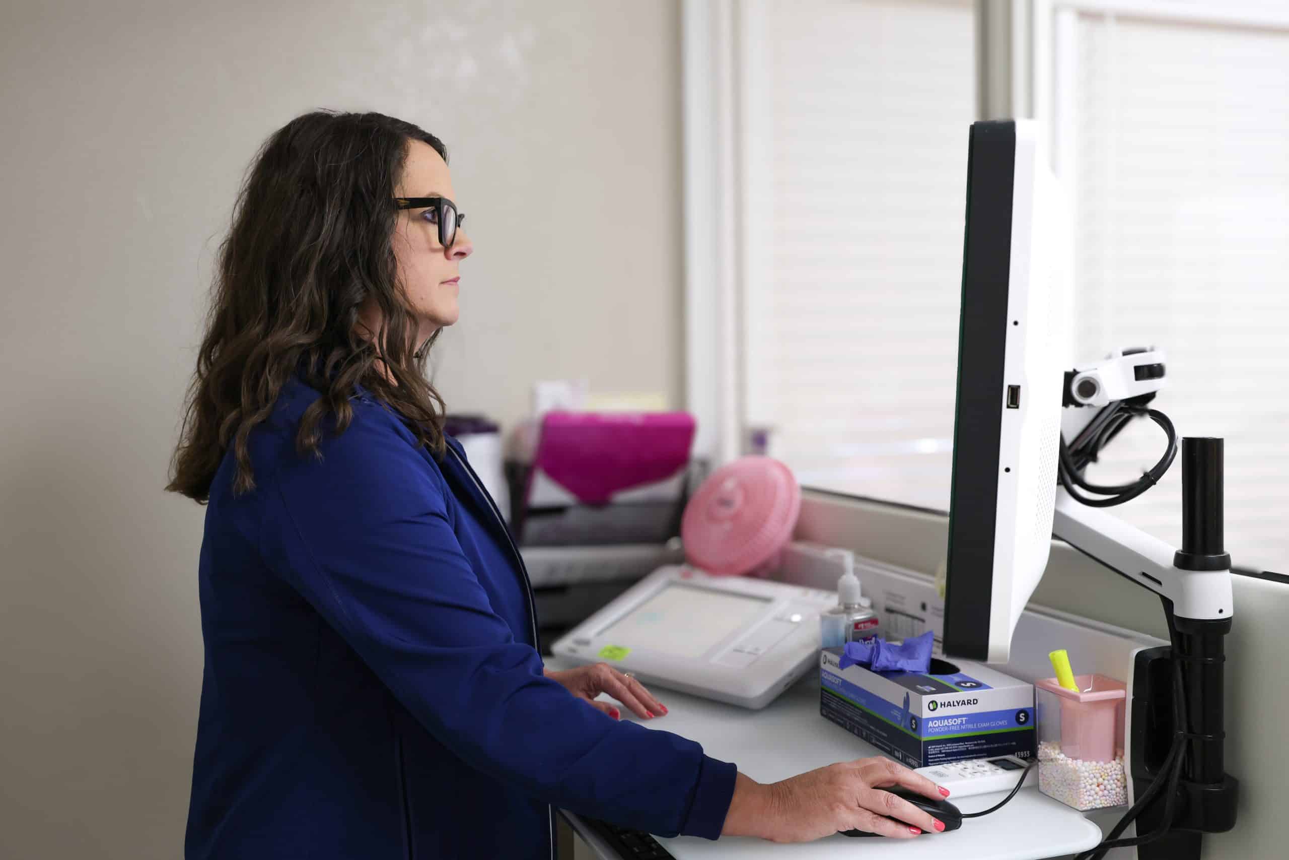 A woman in blue jacket working at a computer station in an office setting.