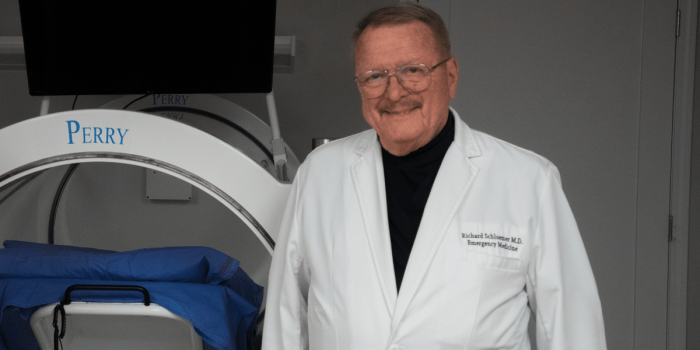 Richard Schlomer, M.D., in a white coat, stands beside medical equipment labeled Perry.