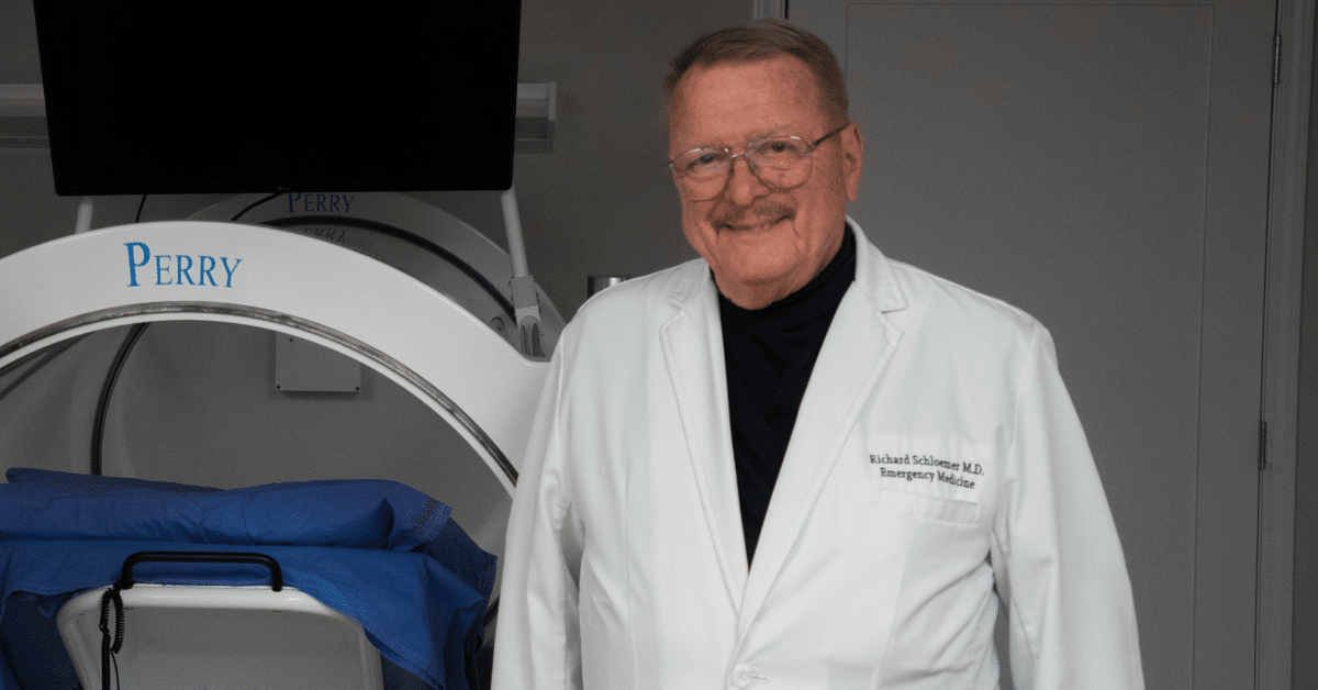 Richard Schlomer, M.D., in a white coat, stands beside medical equipment labeled Perry.
