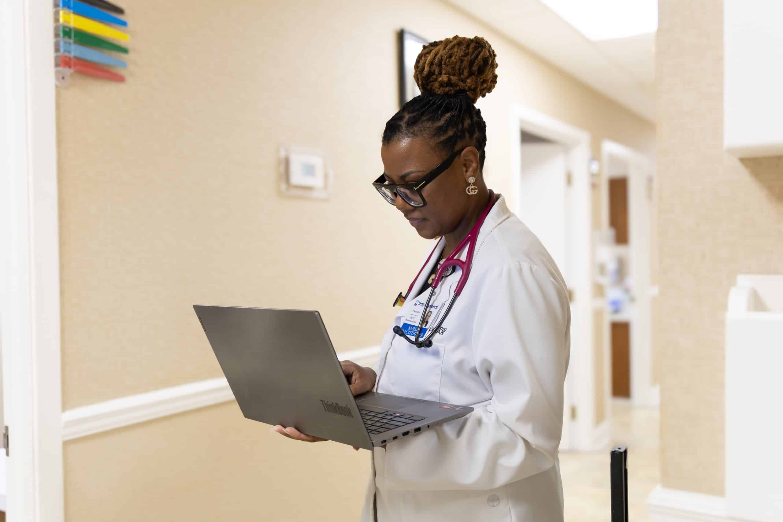 A doctor with a stethoscope working on a laptop in a hospital hallway.