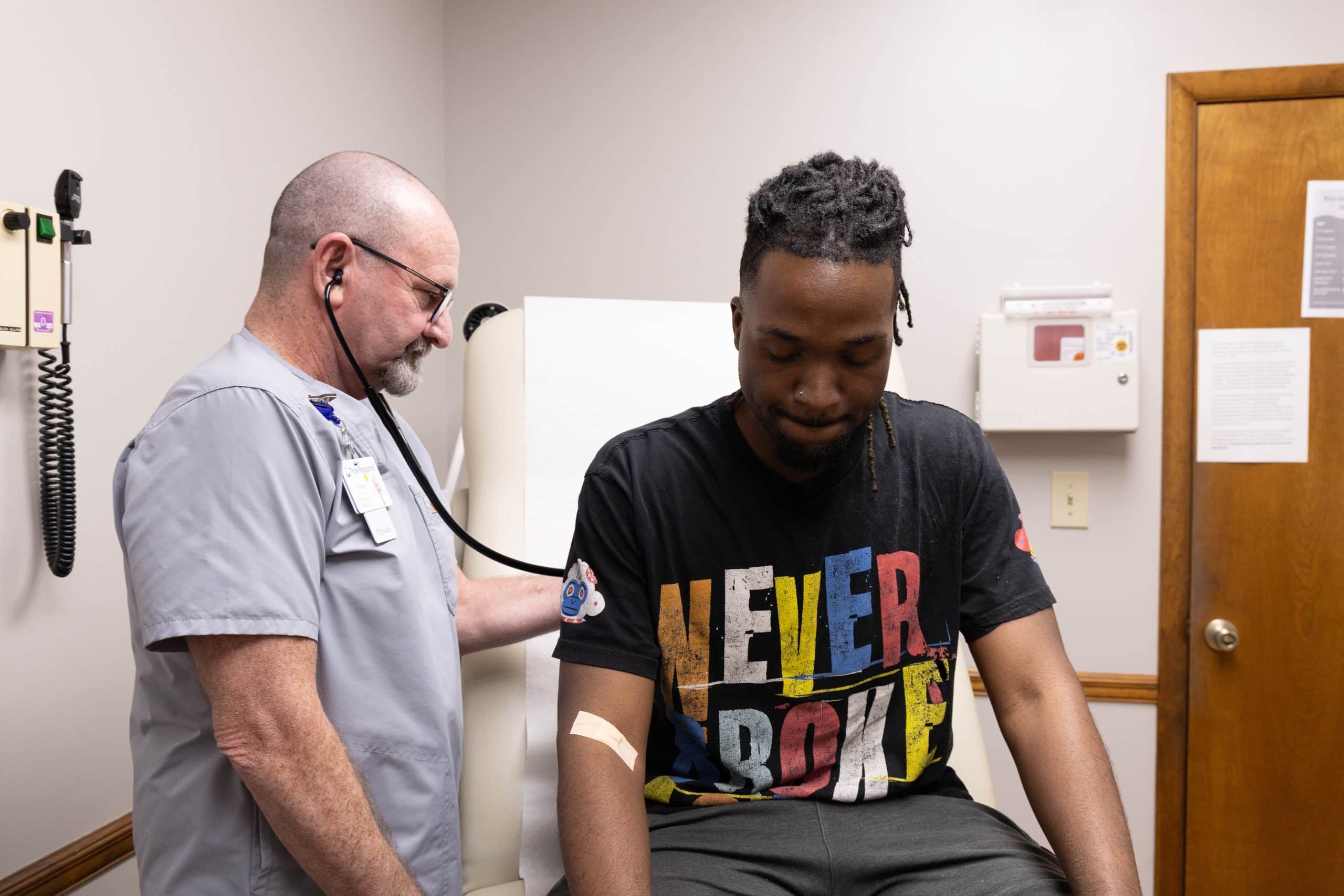 Healthcare professional using a stethoscope on a patient in a clinic exam room.