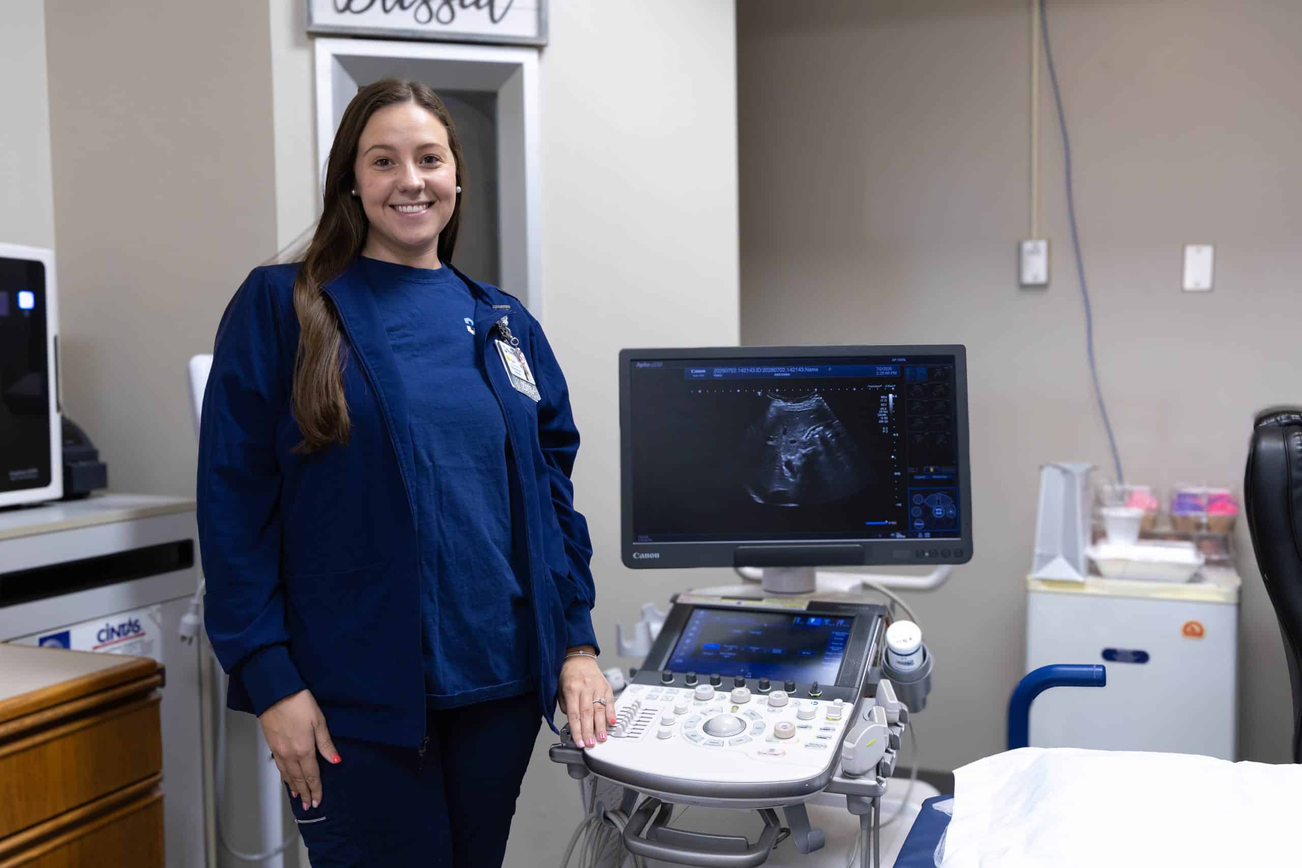 A woman in medical scrubs stands next to an ultrasound machine in a clinic room.