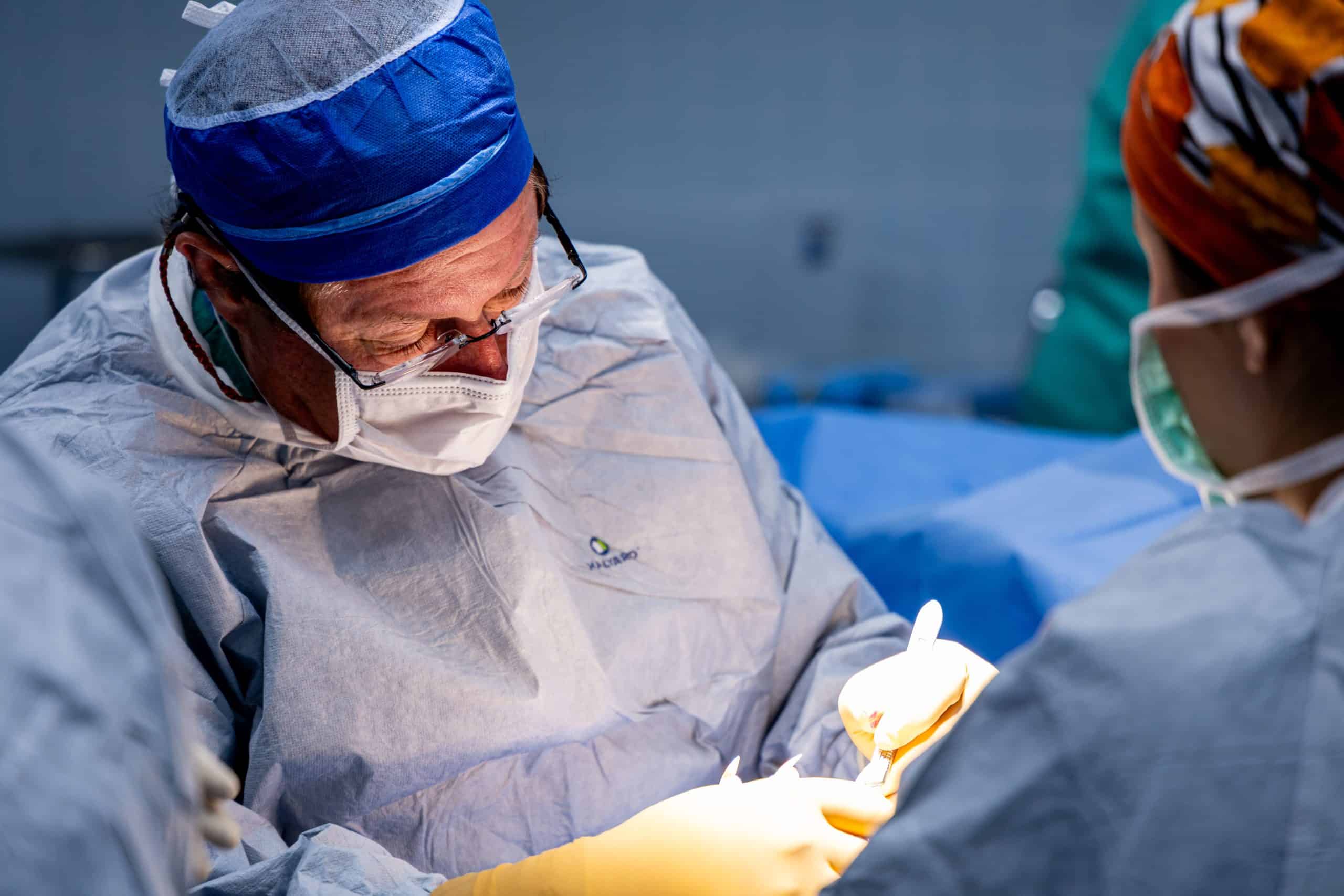 Surgeon in blue cap and mask performing surgery in an operating room, focused and illuminated by surgical lights.